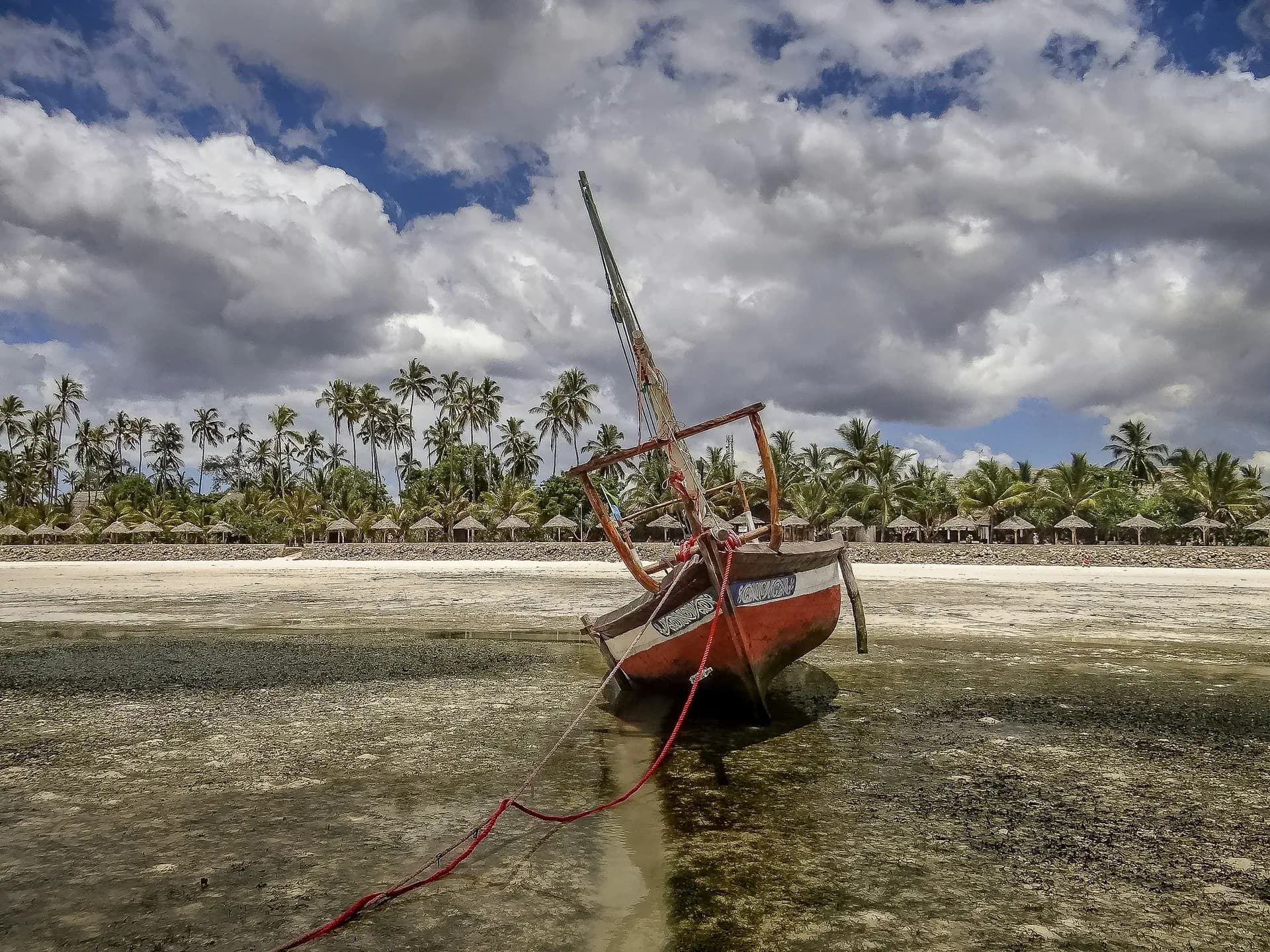 A red-hulled dhow resting on the sand at low tide, palms and thatched umbrellas along the shore behind.