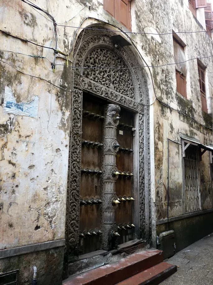 A carved wooden Zanzibar door with brass studs, set in an arched frame on a weathered Stone Town facade.